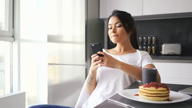 Happy asian woman sitting by the table on kitchen with laptop computer and pancakes while using smartphone