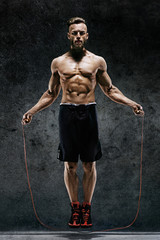 Handsome young man training with his skipping rope. Photo of sporty muscular man with perfect body on dark background. Best cardio workout - obrazy, fototapety, plakaty
