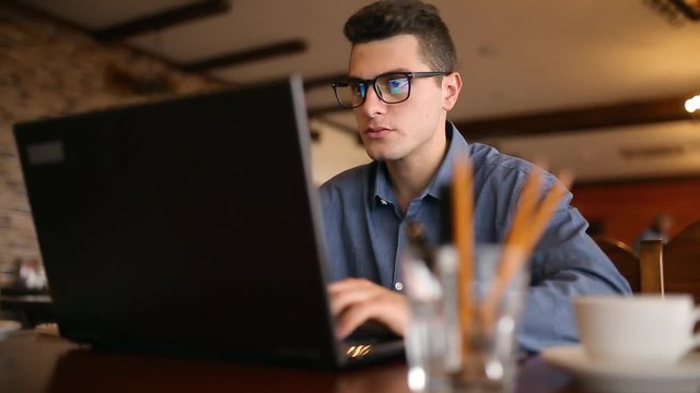 Handsome freelancer businessman in glasses diligently working on laptop in cafe. Man typing on keyboard and searches new job on internet at coffeeshop. Business concept