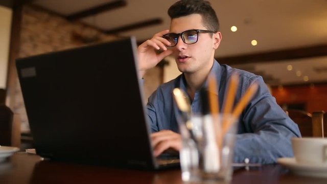Focused young tech guy freelancer interrupts typing on his laptop, removes glasses, smiling and looks dIrectly into the camera. Business concept.