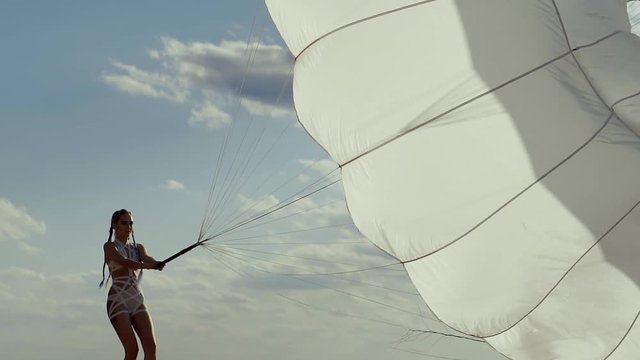 a young woman in modern and urban clothing drags a sail from a parachute to a sandy beach, in the background a clear sky with clouds, a lady with a stylish hairstyle makes an effort