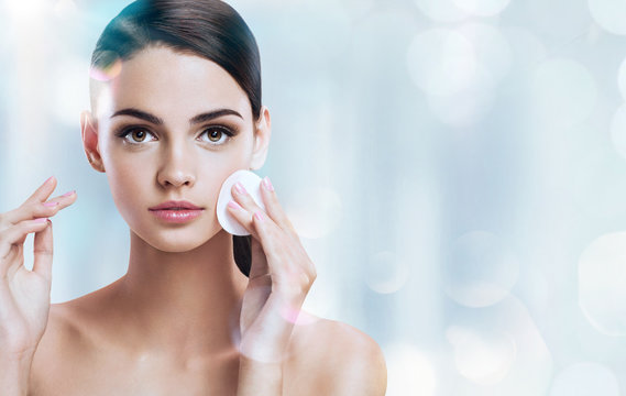Young brunette girl removing makeup from her face. Photo of beautiful girl with cotton pads  on shiny silver background. Skin care and beauty concept.