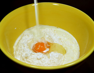 Milk pouring into raw flour and fresh egg in yellow mixing bowl on black background 
