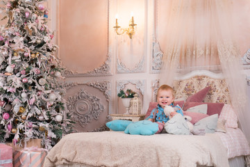 Little happy boy sits on a bed in a pink room with a Christmas tree