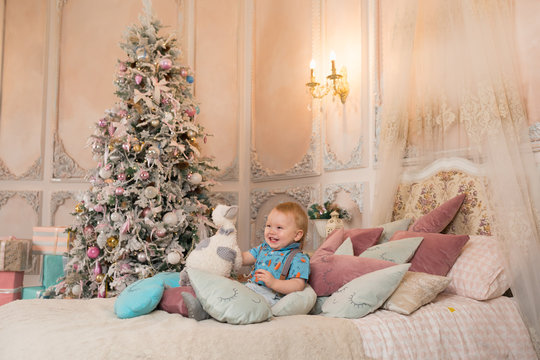 Little happy boy enjoying holidays sitting on a bed in a pink room with a Christmas tree