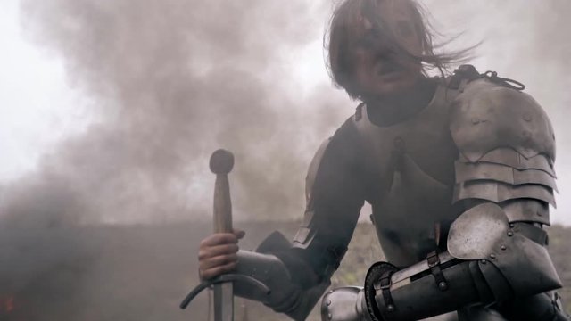 Wounded woman knight sits in smoke and leans on sword, slow motion