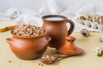 Freshly prepared buckwheat porridge in a small clay pot, next to a mug with milk and quail eggs on an old wooden table. Healthy, natural food. 