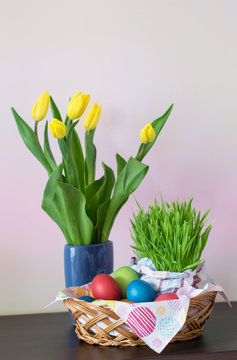 A wicker basket with colorful Easter eggs, yellow tulips in vase, Easter decoration on light background. Free space for your text. Vertical image.