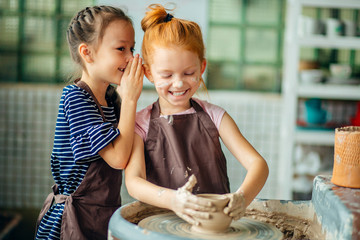pottery workshop - two girls sculpts from clay. one child whispers from her friend
