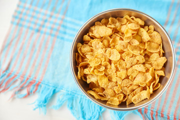 Cornflakes in a metal bowl on a painted white wooden background. Composition with a dish towel.