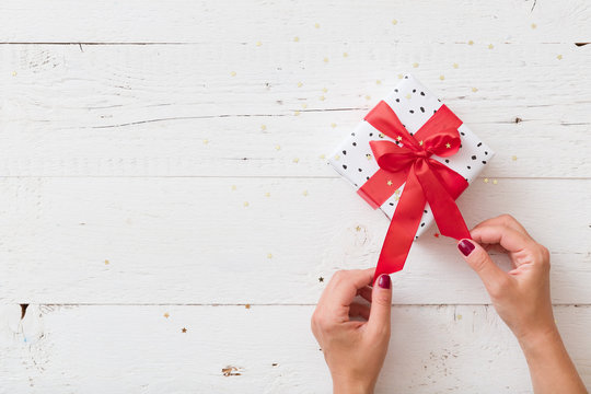 Top View On Woman's Hands Unwrapping Christmas Or Birthday Gift Box With Red Satin Ribbon On White Wooden Background.