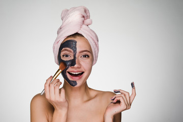 Laughing beautiful girl with a pink towel on her head puts on half face clay mask