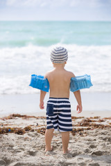 Back view on cute toddler boy wearing safety inflatable armbands standing on the beach ready to...