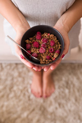 Top view on girl's hands holding black bowl with organic granola with coconut milk and raspberries.