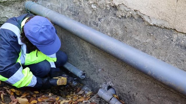 A woman conducts plumbing in difficult conditions in a narrow pit. The drain pipe burst under the foundation of the house. To replace it, the moman plumber drill a hole for fastening.
