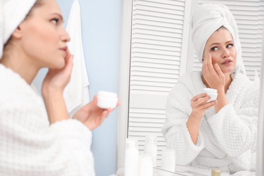 Young woman in towel and bathrobe using cosmetic product at home