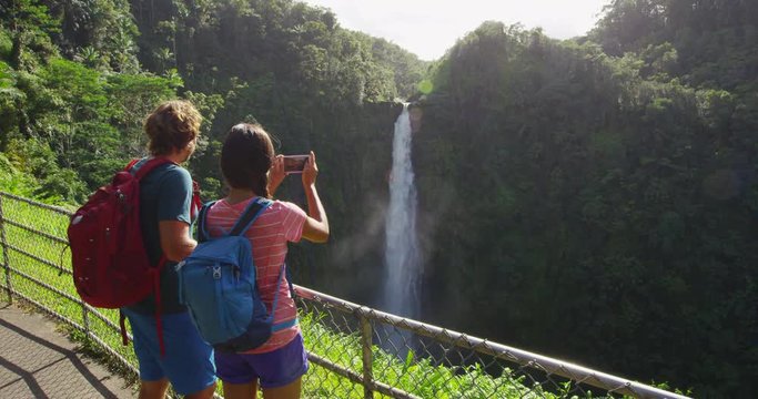 Couple tourists on Hawaii by waterfall. Tourist taking photo using phone of Akaka Falls waterfall on Hawaii, Big Island, USA. Travel tourism concept with multicultural tourist couple.
