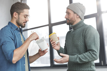 Photo of professional journalists meet together at office for discussing creating new article, eat junk food and drink coffee from paper cup, communicate during work break, stand over modern interior