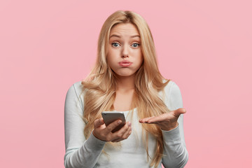 Indoor shot of indignant puzzled woman blows cheeks as looks into screen of modern cell phone, can`t understand content of recieved message, has frustrated look. Technology and people concept