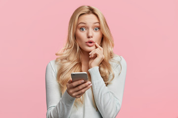 Beautiful amazed female model recieves text message on mobile phone, shocked get reminder to pay her bills being puzzled, isolated over pink background. Surprisment and modern technology concept