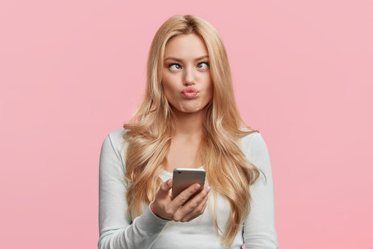 Crazy Funny Comic Female Crosses Eyes, Pouts Lips, Makes Grimace, Foolishes, Holds Cell Phone, Involved In Games Online, Isolated Over Pink Background. Facial Expressions. Childish Woman Plays Fool