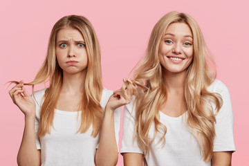 Isolated shot of cheerful blonde female and her companion blows cheeks as touches hair, being dissatisfied with her new shampoo, isolated over pink background. Two friends meet together, gossip
