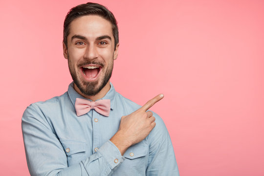 Handsome brunet young male with dark beard and mustache dressed in trendy shirt and bowtie, indicates with excited expression at blank pink copy space for your advertising or promotional text