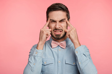 Brunet discontent handsome young male model with trendy hairdo frowns face in displeasure, being forgettable, fails to concentrate as has terrible headache after conference, dressed formally