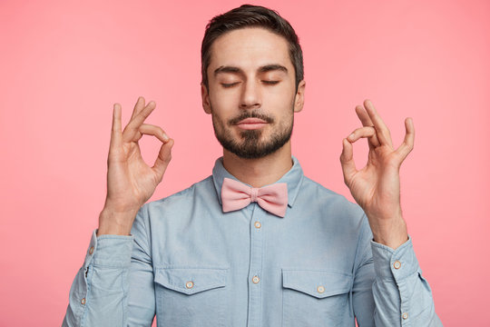 Calm peaceful atmosphere. Relaxed attractive male model meditates and tries to relax for minute, keeps eyes shut, stands in mudra gesture, dreams about something, isolated over pink background