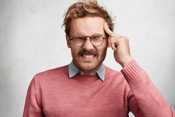 Horizontal shot of fashionable trendy looking stressful male model clenches teeth, keeps eyes shut,...