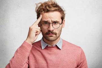 Muurstickers Portrait of handsome young stylish male tries to concentrate as sees something in front, has problematic expression, dressed in pink sweater, has round eyewear, isolated over white concrete wall © Wayhome Studio