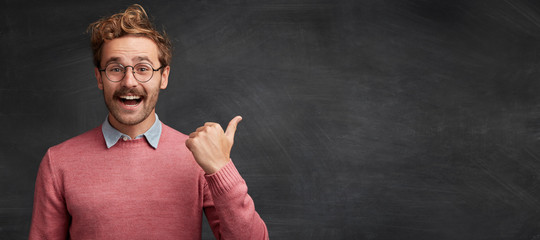 Horizontal shot of bearded fashionable male student indicates at blank chalk blackboard with thumb,...