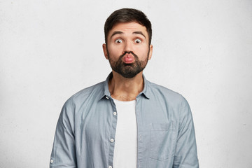 Funny unshaven male makes grimace, pouts lips and stares at camera, has comic expression, dressed in fashionable clothes, stands alone against white studio background. Facial expressions concept