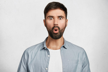 Obraz premium Amazed bearded shocked man expresses great surprisment, dressed in fashionable clothes, being astonished to be promoted, poses against white background. Stunned unshaven male model in studio
