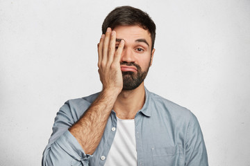 Bearded male brunet covers face as shows his tiredness, curves lips, has no desire to work, dressed in denim shirt, isolated over white concrete background. Frustrated man with indignant look