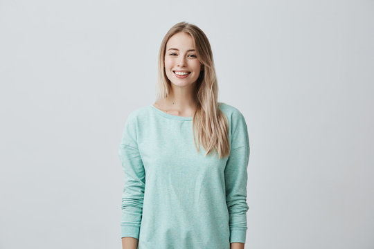 European female model with long blonde hair, wears light blue sweater, looks at camera with dreamful and positive expression imagines or remembers pleasant moments spent with boyfriend.