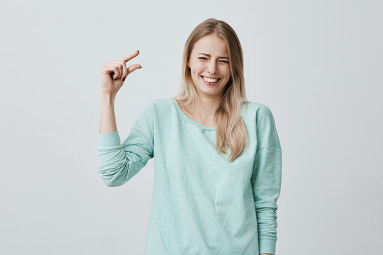 Pretty cute blonde girl in casual clothes showing something small in size with hands while gesturing, smiling broadly with teeth. Blonde European female demonstrating size of little box