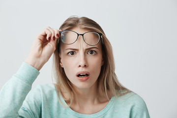 Surprised young female model with long blonde hair, wears glasses and blue long-sleeved shirt,...