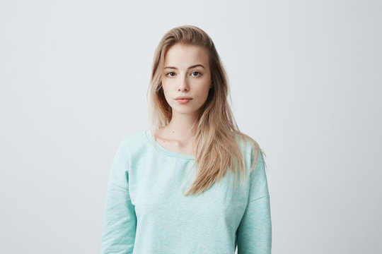People and lifestyle. Studio shot of attractive young Caucasian dark-eyed female with long dyed blonde hair posing against gray blank wall dressed in casual blue sweater with calm face expression.