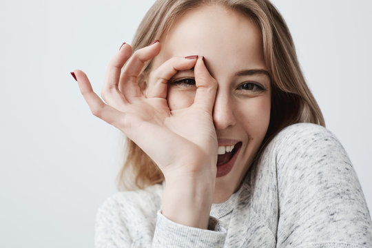 Young European joyful blonde female looking at camera through fingers in OK-gesture. Woman in casual clothes, smiling broadly. Her happy face expression proves everything goes according to plan.