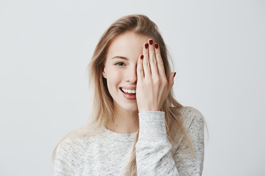 Happy smiling female with attractive appearance and blonde hair wearing loose sweater showing her broad smile having good mood closing her eye with hand, enoying to pose at camera. Happiness and joy