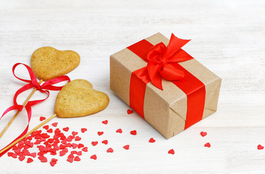 Box with a gift wrapped in kraft paper and tied with a red ribbon and two homemade cookies on a stick in the shape of hearts on white wooden table. Valentines Day concept.