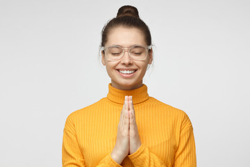 Closeup of attractive young female in transparent glasses putting hands together as if she is praying with closed eyes, smiling