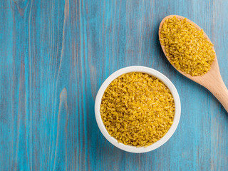White bowl  and wooden spoon with raw bulgur on blue wooden background. Top view, copy space for text.