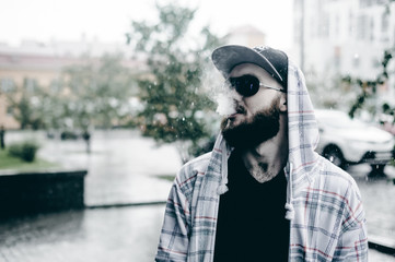 young man with beard in light checkered jacket with hood in a cap and sunglasses walking freely around city smokes an e-cigarette - Powered by Adobe