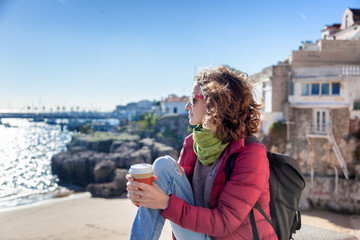beautiful young woman student tourist with a glass of coffee in hands, against the backdrop of the coast of a European city, sun, wind, freedom, spring
