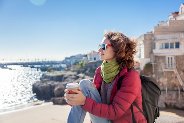 Fototapeta na wymiar beautiful young woman student tourist with a glass of coffee in hands, against the backdrop of the coast of a European city, sun, wind, freedom, spring