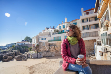 Fototapeta na wymiar beautiful young woman student tourist with a glass of coffee in hands, against the backdrop of the coast of a European city, sun, wind, freedom, spring