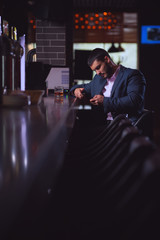 Side view - young handsome man in formal attire is drinking whiskey in bar and writing message on smartphone inviting friends to join