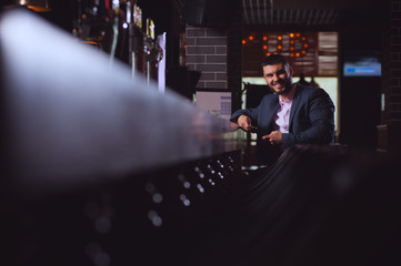 Positive handsome man businessman rests in bar after  long working day and holding smartphone in hands answering messages in social networks